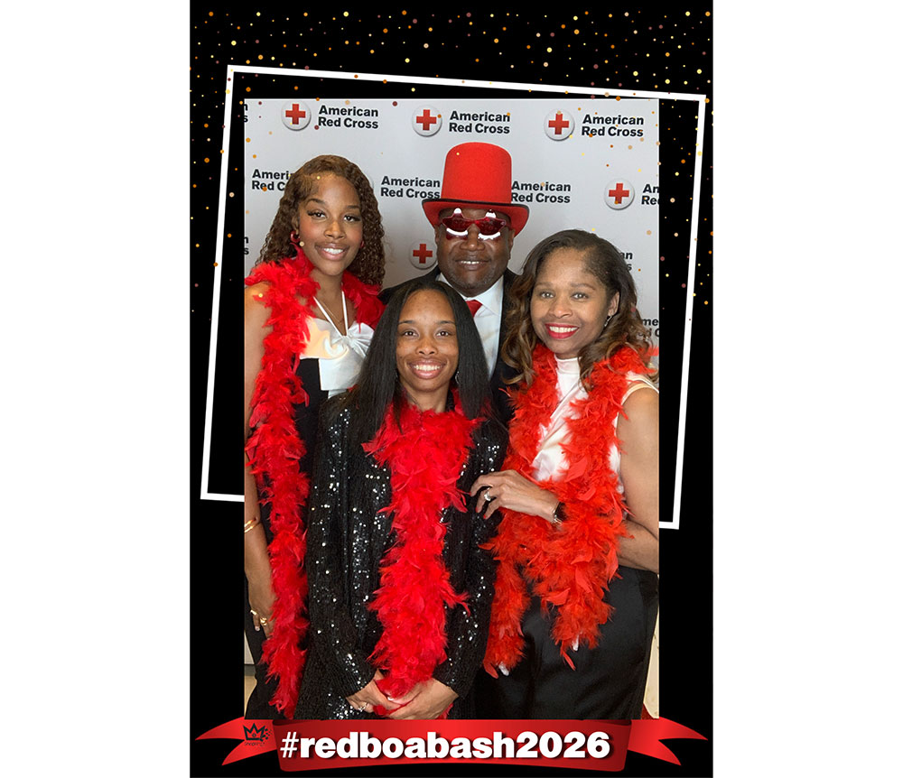 A group of four people smile in front of an American Red Cross backdrop, wearing red feather boas. The mood is festive, with the hashtag #redboabash2026.