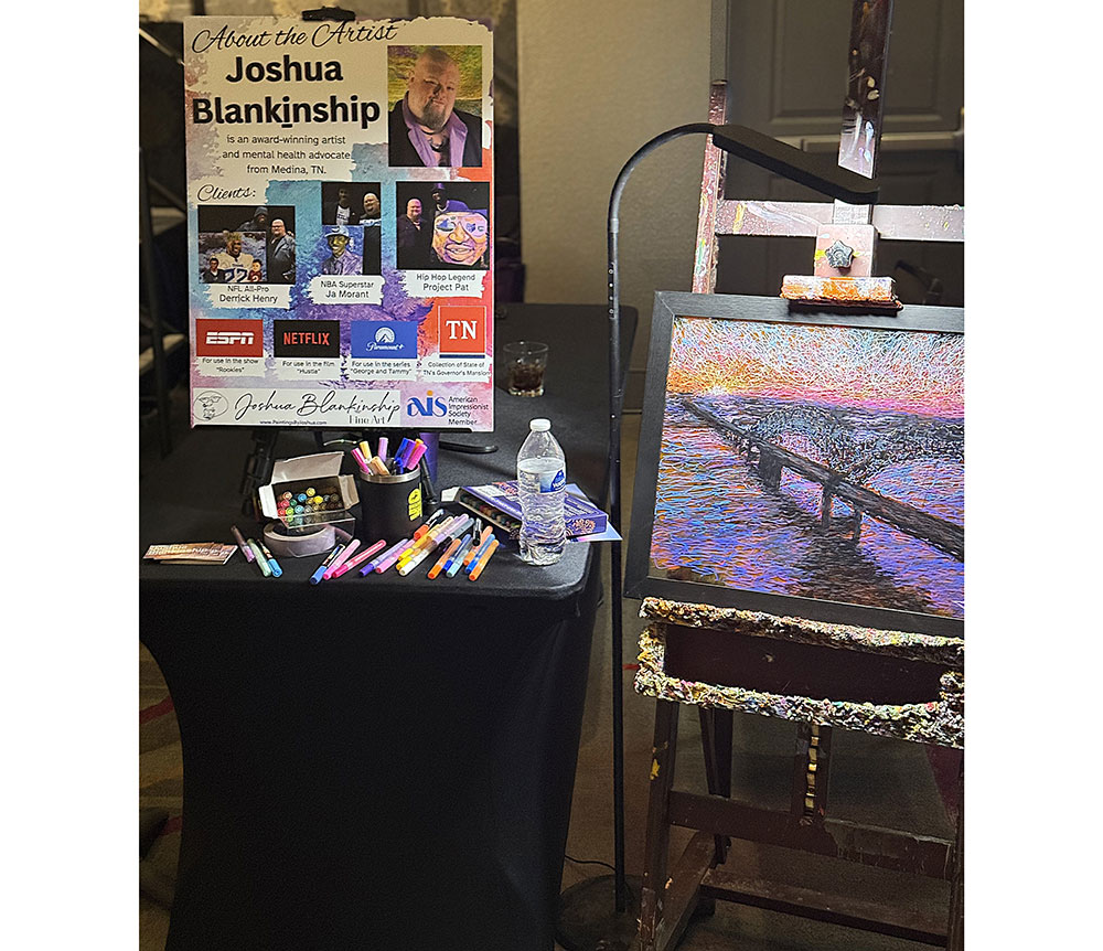 Art display featuring an information board for artist Joshua Blankinship. A colorful painting of a bridge is on an easel, with art supplies on a table. The scene is vibrant and creative.