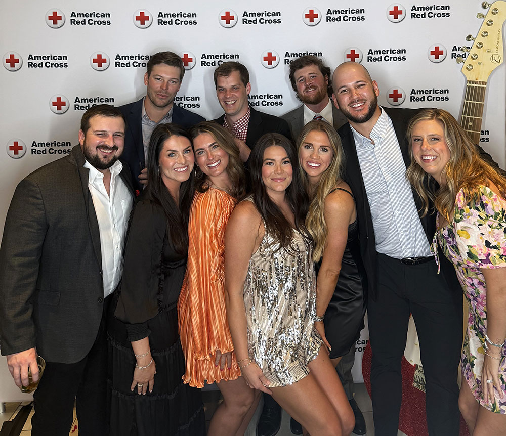 A group of people smiling in front of a backdrop with "American Red Cross" logos. The atmosphere is joyful and festive, suggesting a celebratory event.