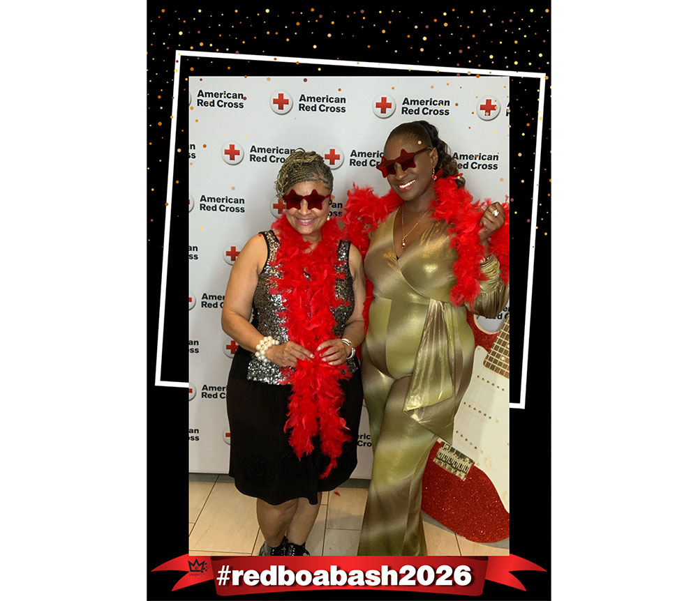 Two people smiling, wearing star-shaped sunglasses and red feather boas, pose in front of an American Red Cross backdrop. Text reads "#redboabash2026."