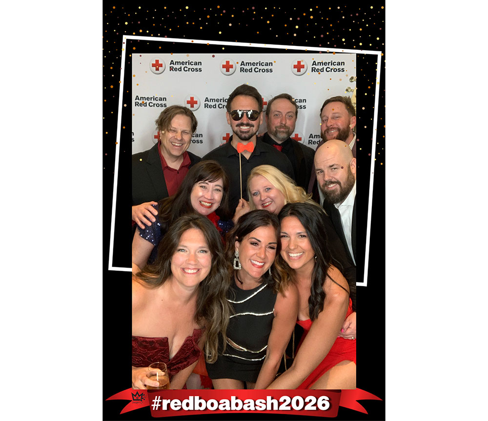 A group of smiling people at an American Red Cross event, dressed in formal attire, with a backdrop featuring the Red Cross logo. The atmosphere is celebratory.