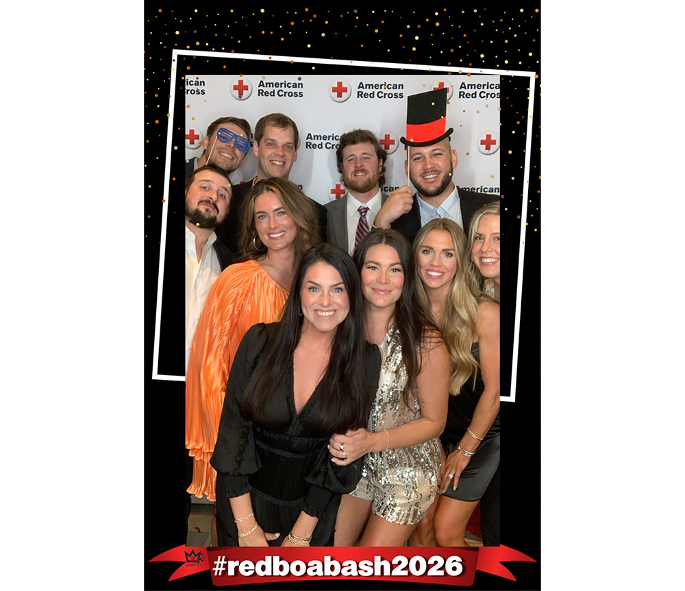 Group of smiling friends posing at a party in front of an American Red Cross backdrop. Some wear formal attire, with one wearing a digital top hat. Hashtag #redboabash2026.