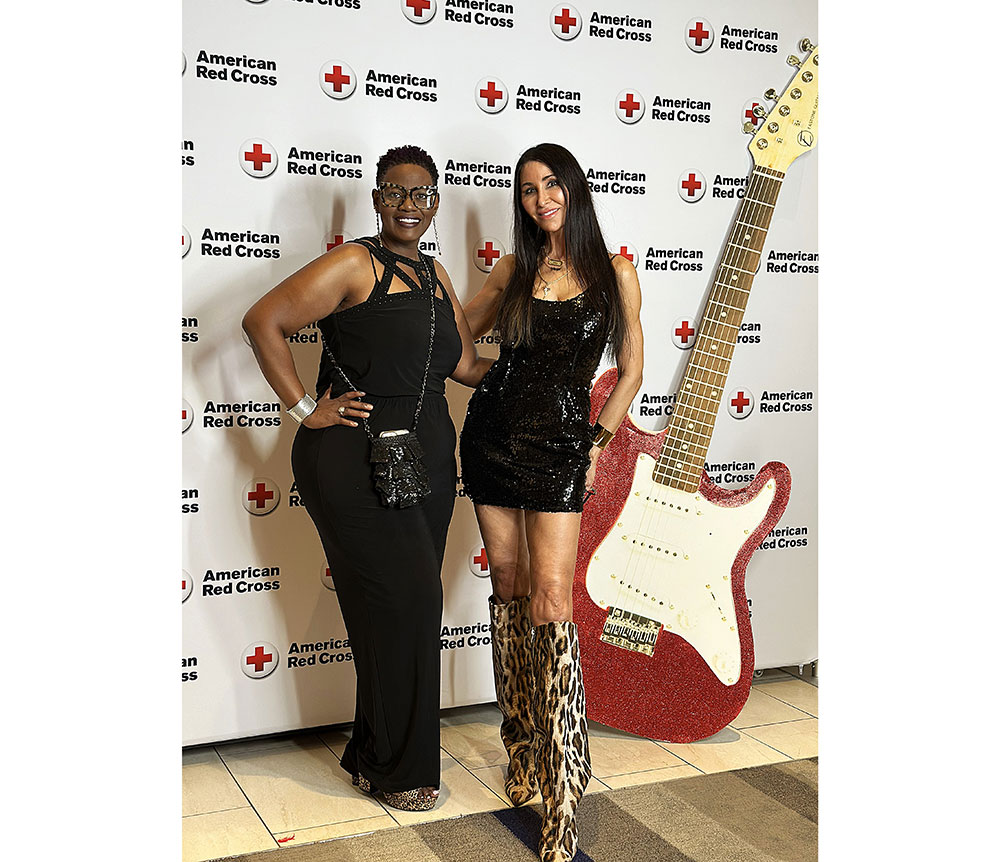 Two individuals pose confidently at an event with an American Red Cross backdrop and large red guitar prop, exuding a celebratory atmosphere.
