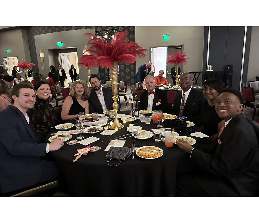 A group of eight people sit around a table at a formal event. The table is decorated with a tall gold centerpiece with red feathers. Plates of food, drinks, and notes are visible. The room is elegant, filled with similar tables. The atmosphere is festive and lively.