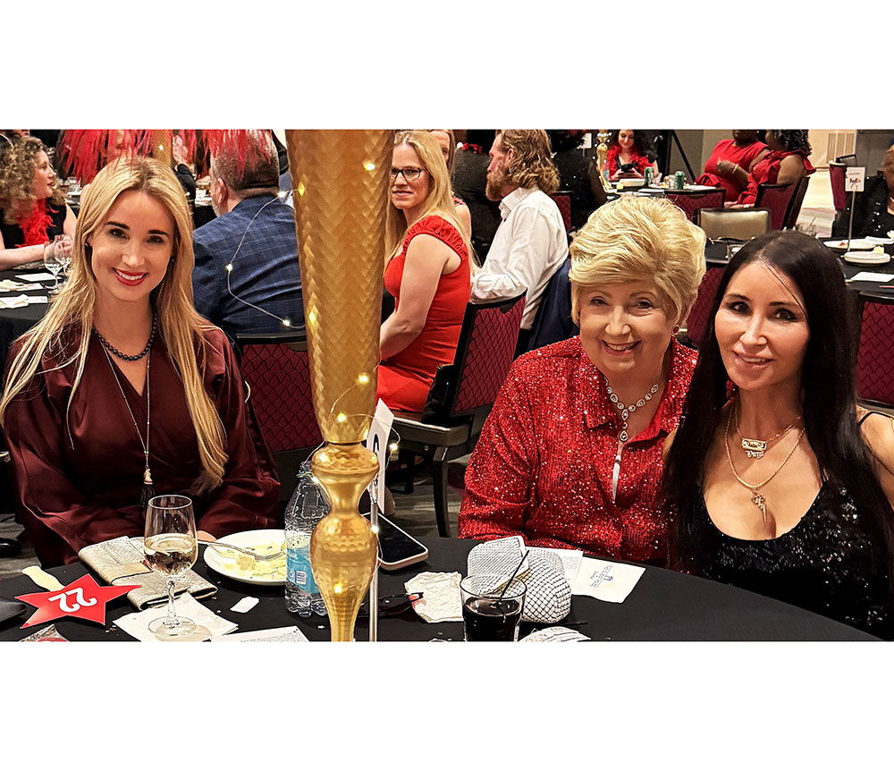 Three women sit at a formal dinner, smiling warmly. They wear elegant dresses in red and black with sparkling details. The atmosphere is festive.