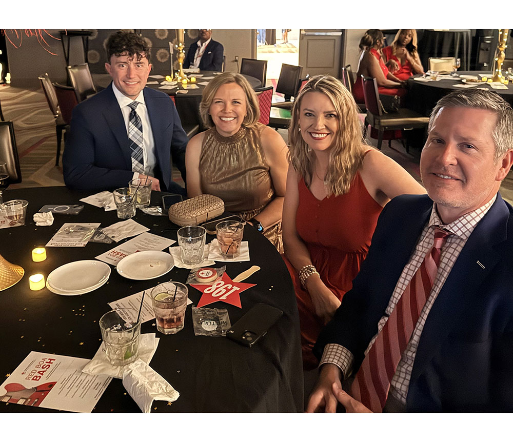 Four people smiling at a gala event, seated around a table with drinks and event materials. The mood is festive and the setting is elegant.