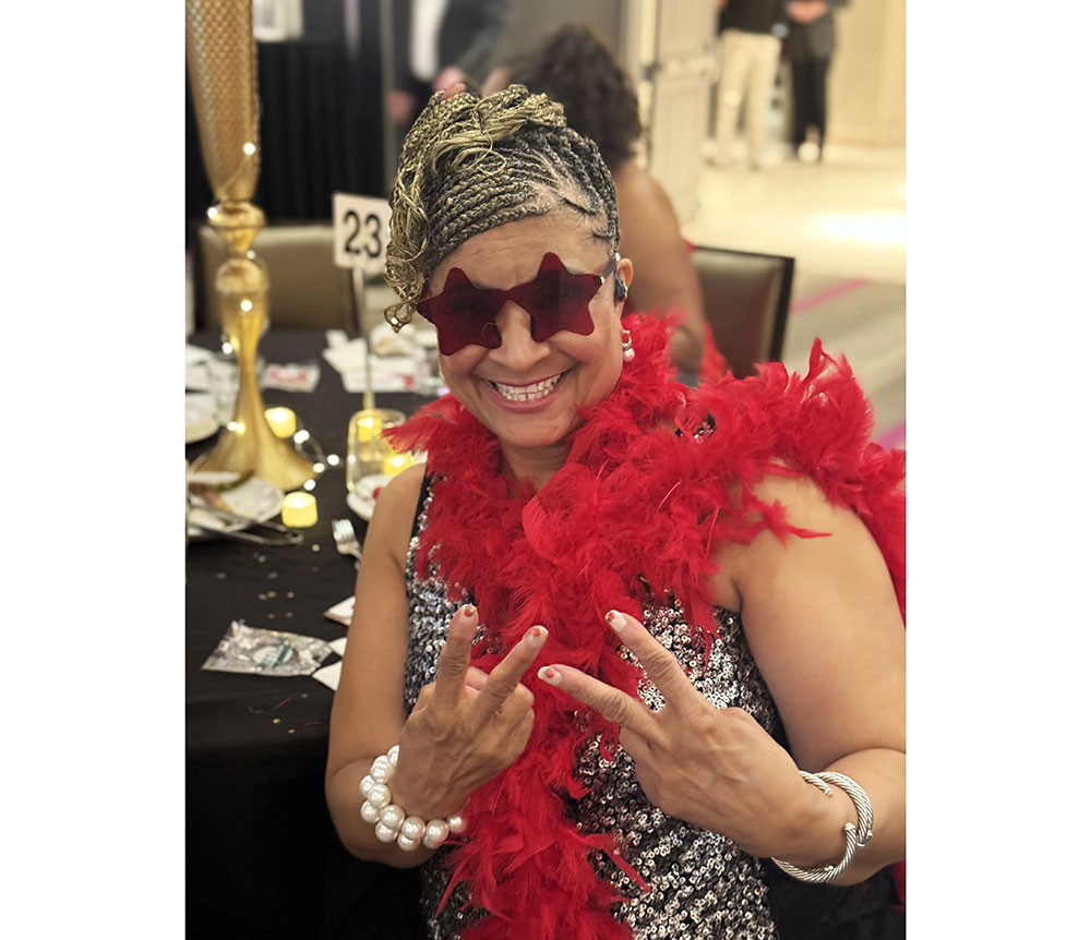 Smiling woman in a sparkly dress and red feather boa wears star-shaped sunglasses, flashing peace signs. Festive setting with decorative tableware.