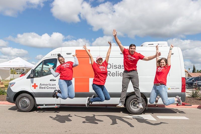 Four Red Cross volunteers jumping and raising hands in excitement with Red Cross van behind them.