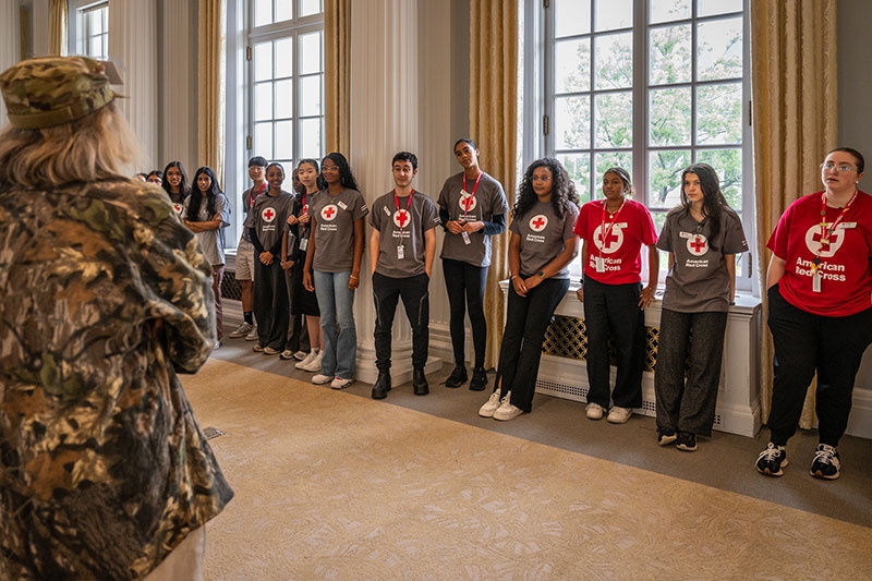 Military memebr speaking to a group of teen Red Cross volunteers in a room.