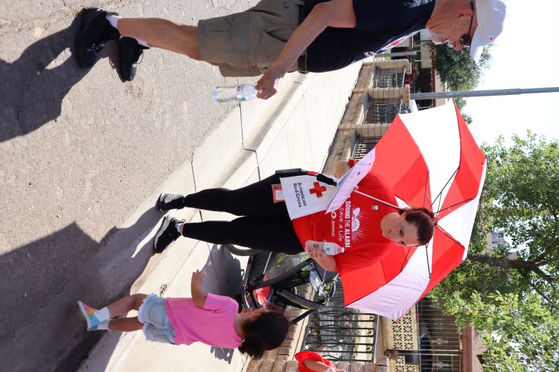 Red Cross volunteer under umbrella handing out a bag to a child.