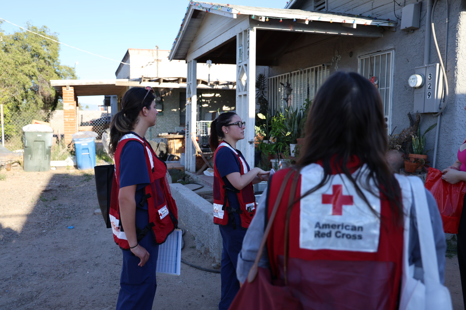 A group of Red Cross disaster relief workers in red vests walk down a sunny street lined with palm trees and parked cars.
