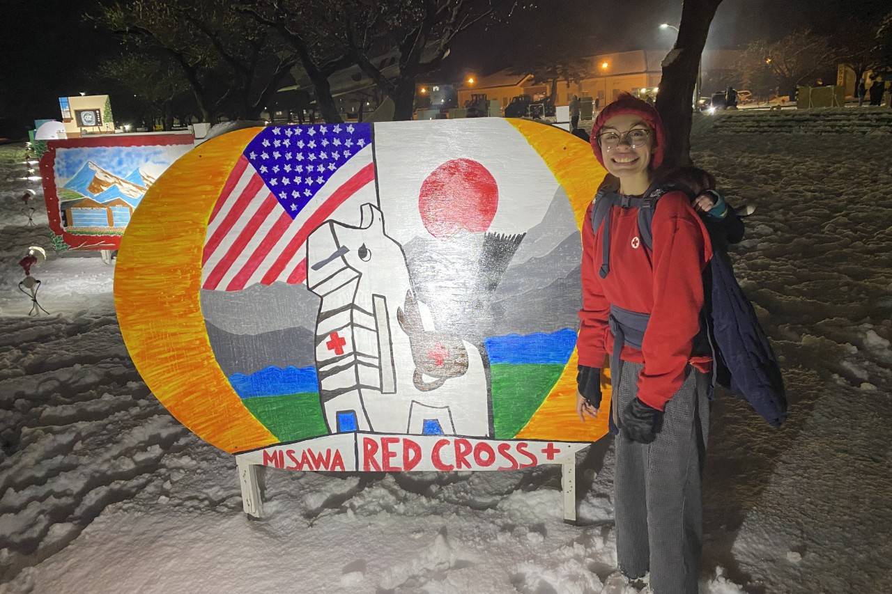 Woman stands in front of wooden painted sign wearing Red Cross gear