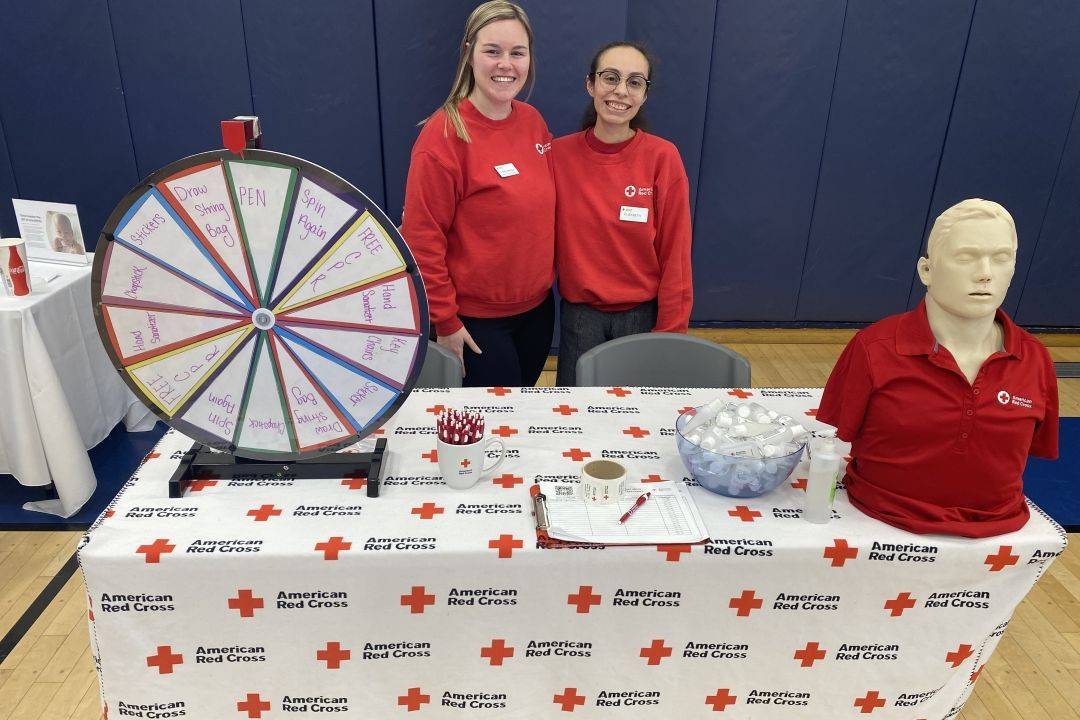 Two women stand behind Red Cross table with spinning wheel