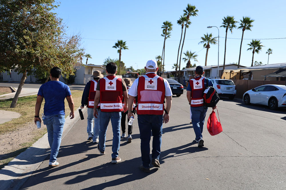 A group of Red Cross disaster relief workers in red vests walk down a sunny street lined with palm trees and parked cars.