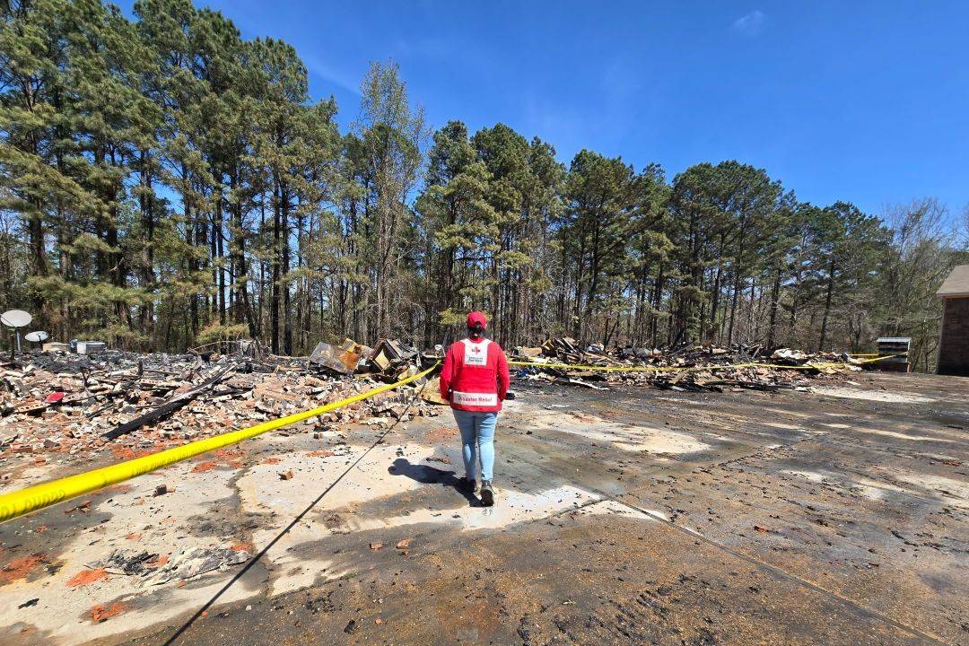 Woman in Red Cross vest walks outdoors among disaster damage