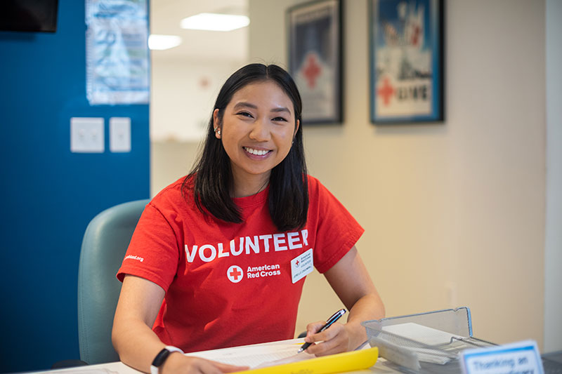 American Red Cross volunteer siting at desk with pen and paper and smiling for picture.