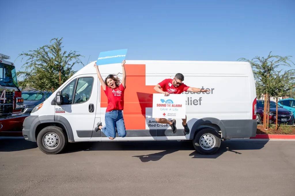 Man and woman jump in front of Red Cross vehicle holding signs