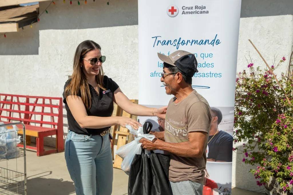 Woman in Red Cross shirt hands bags of items to man 