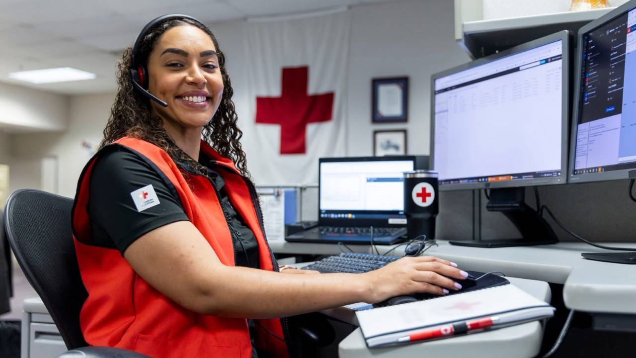 A Red Cross volunteer sitting at a desk looking at a computer with headphones on.