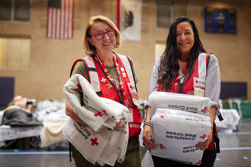 Two Red Cross volunteers holding folded Red Cross blankets in a Red Cross shelter.