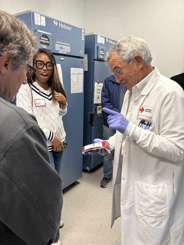 Man in white lab coat holds bag of donated blood while talking with group