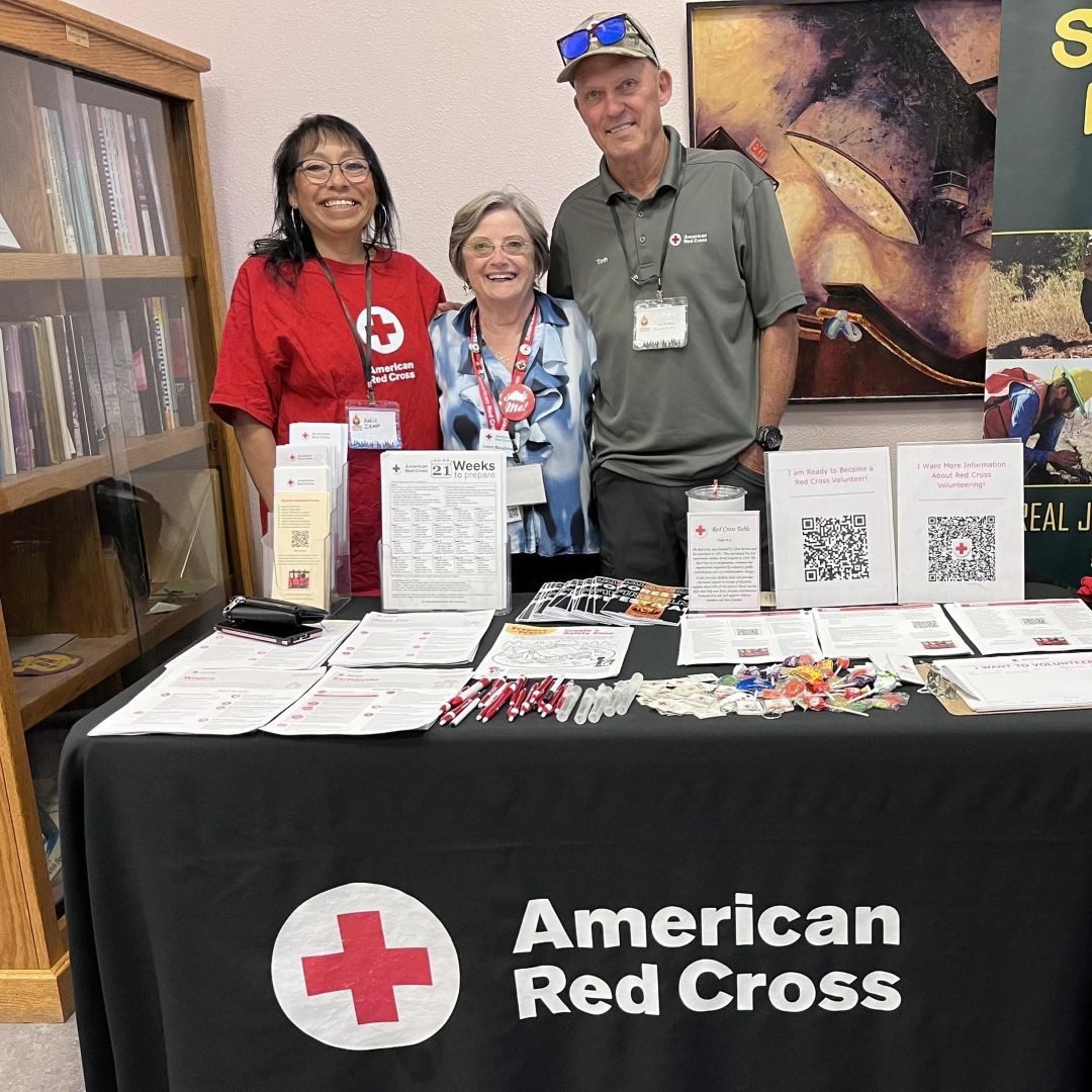 three people stand behind table with red cross logo and resources