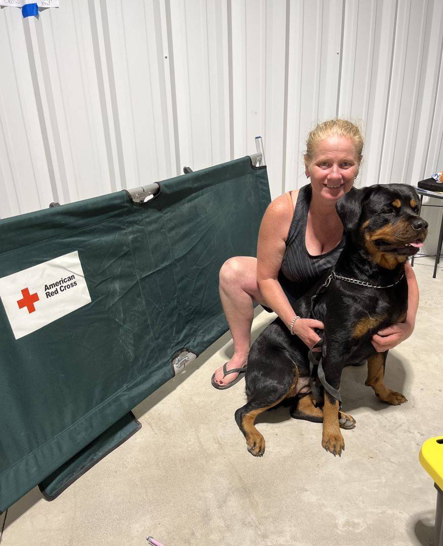 Woman sits in front of Red Cross cot with arms around her dog