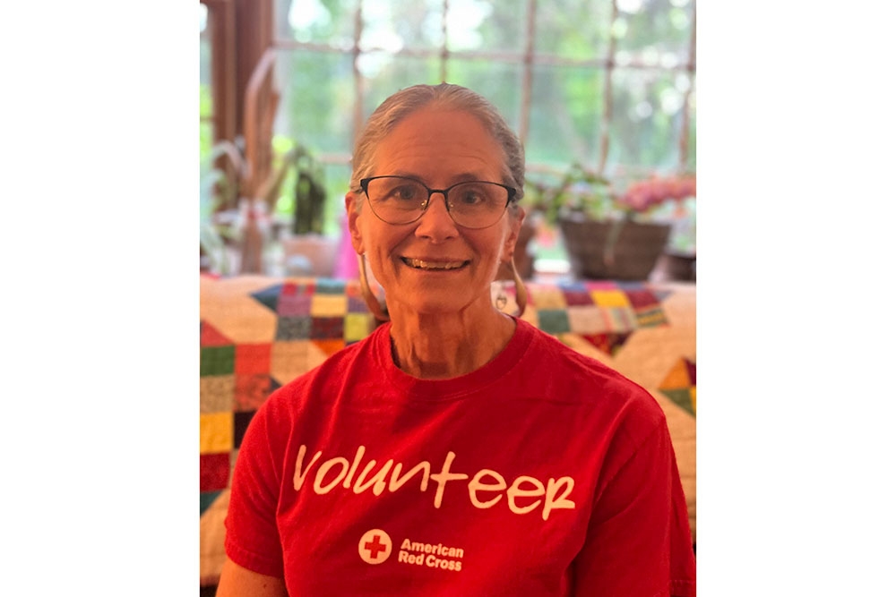 Anne O’Neill smiling wearing glasses and a Red Cross volunteer shirt.