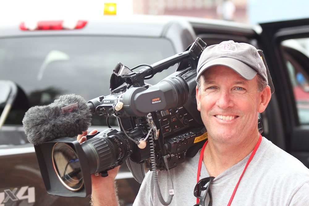 David Zapatka smiling wearing a hat and holding a news camera on his shoulder.