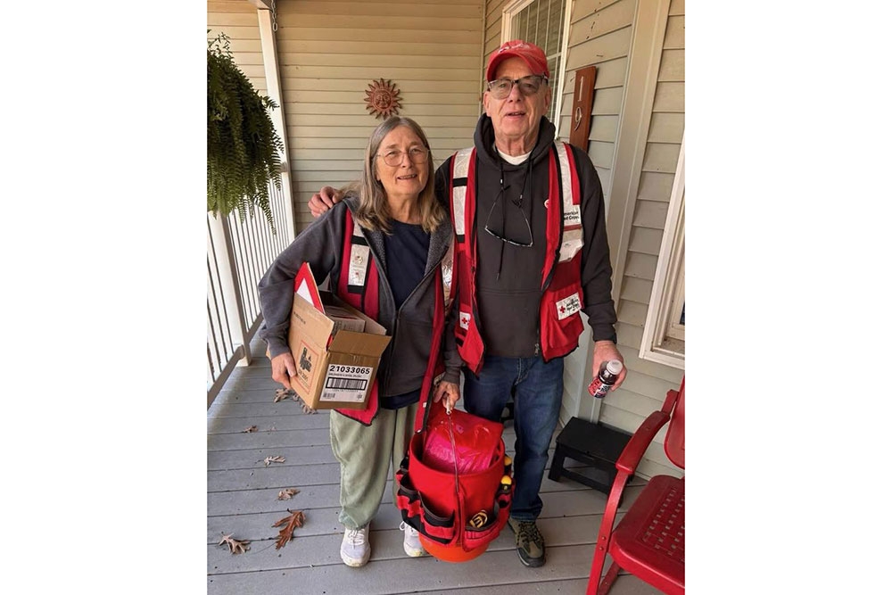 two Red Cross volunteers standing on a house porch and holding a box and tools.