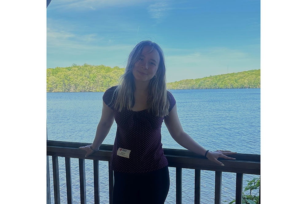 Gabby Remiszewski leaning on a wooden rail with a lake, mountains and a blue sky in the backround.
