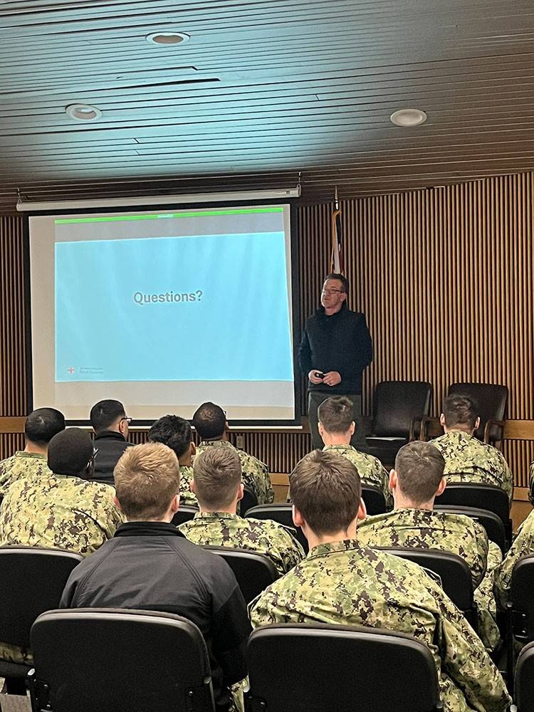 Joe Graziano stands in front of a seated group of military personnel in camouflage uniforms. A slide behind him reads "Questions?" in a classroom setting.