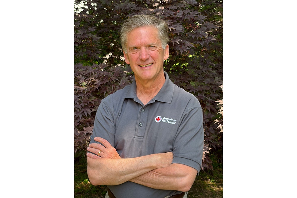 Ken Sneider smiling wearing a Red Cross shirt with his arms folded.