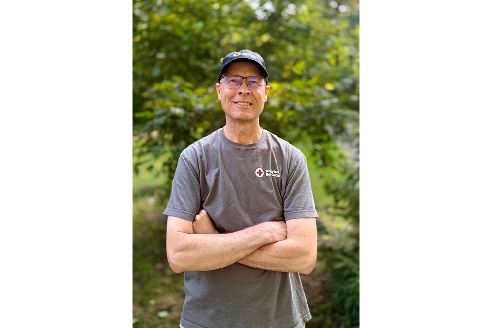Scott Lee wearing a Red Cross hat and shirt stands smiling with his arms folded.