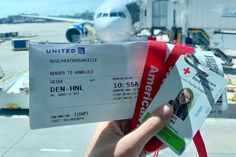 Hand holding airplane ticket, Red Cross badge in front of window with jet in background