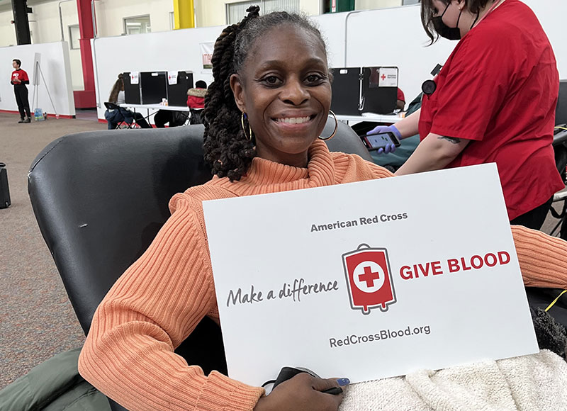 Blood donar sitting in cot holding a give blood sign and smiling for picture.