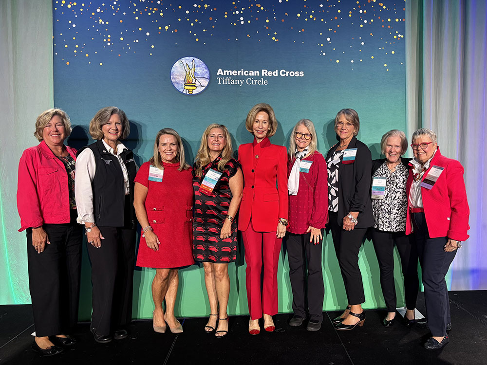 Nine Tiffany Circle members in business clothing and wearing name tags take a group photo next to a large American Red Cross Tiffany Circle logo.