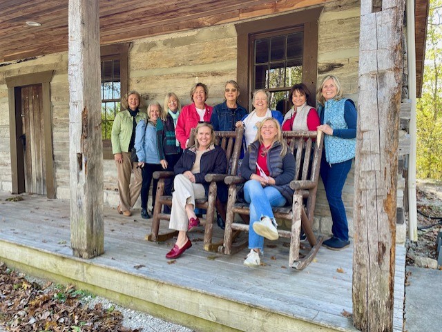 Group of women standing in front of a old cottage with two women sitting in rocking chairs.