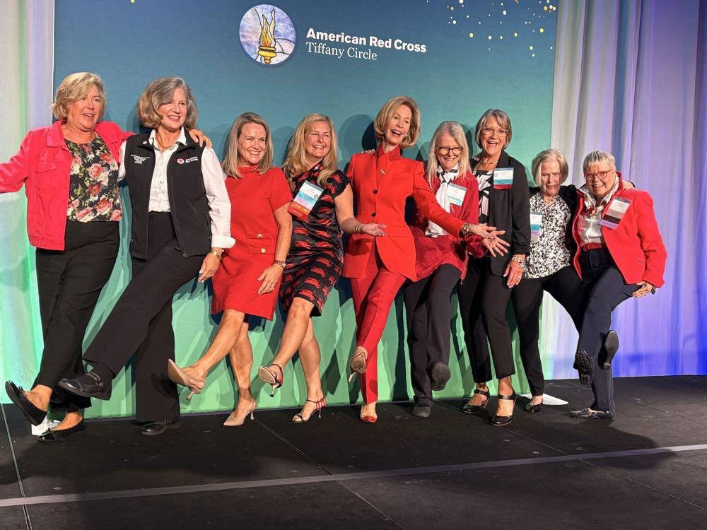 Nine Tiffany Circle members in business clothing, wearing name tags, and doing a leg kick take a group photo next to a large American Red Cross Tiffany Circle logo.