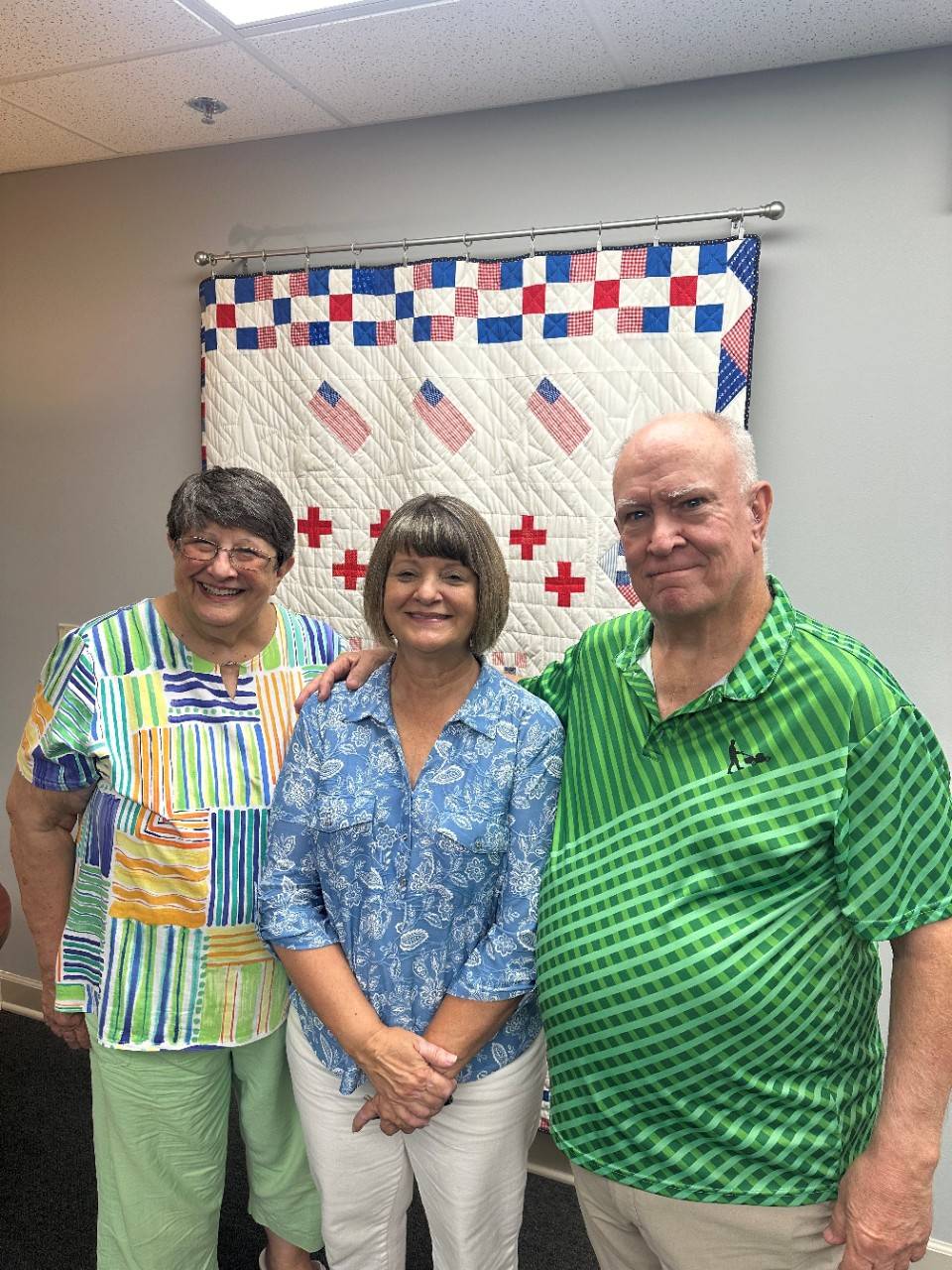 Two women and one man stand in front of Red Cross quilt