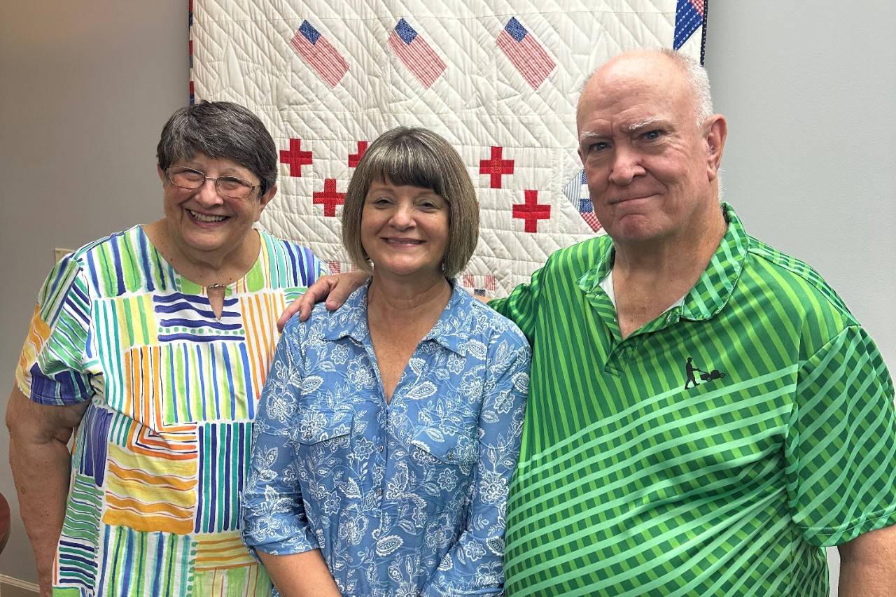 Two women and one man stand in front of Red Cross quilt