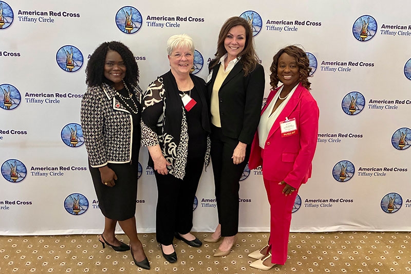 Four women stand smiling in front of an American Red Cross Tiffany Circle backdrop. They are dressed in professional attire.