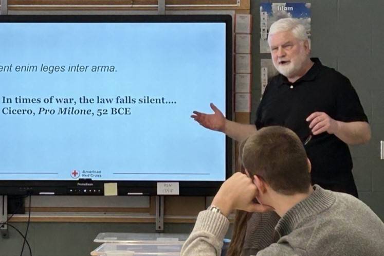 Man in black polo stands in front of screen and speaks to room of people