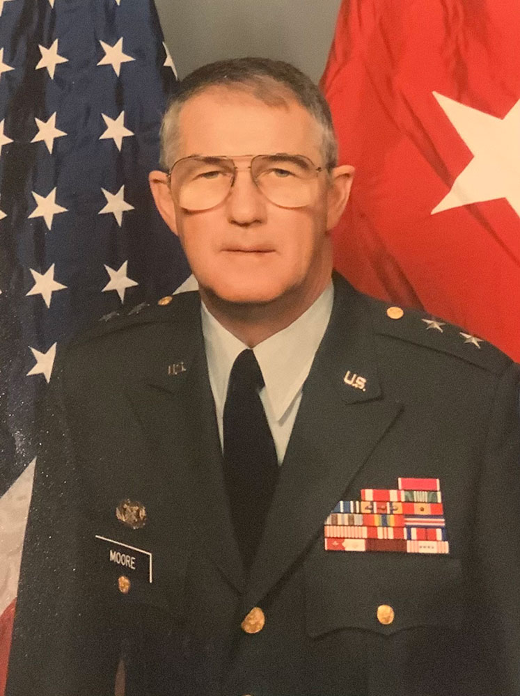 Buddy Moore in military uniform stands proudly beside an American flag.