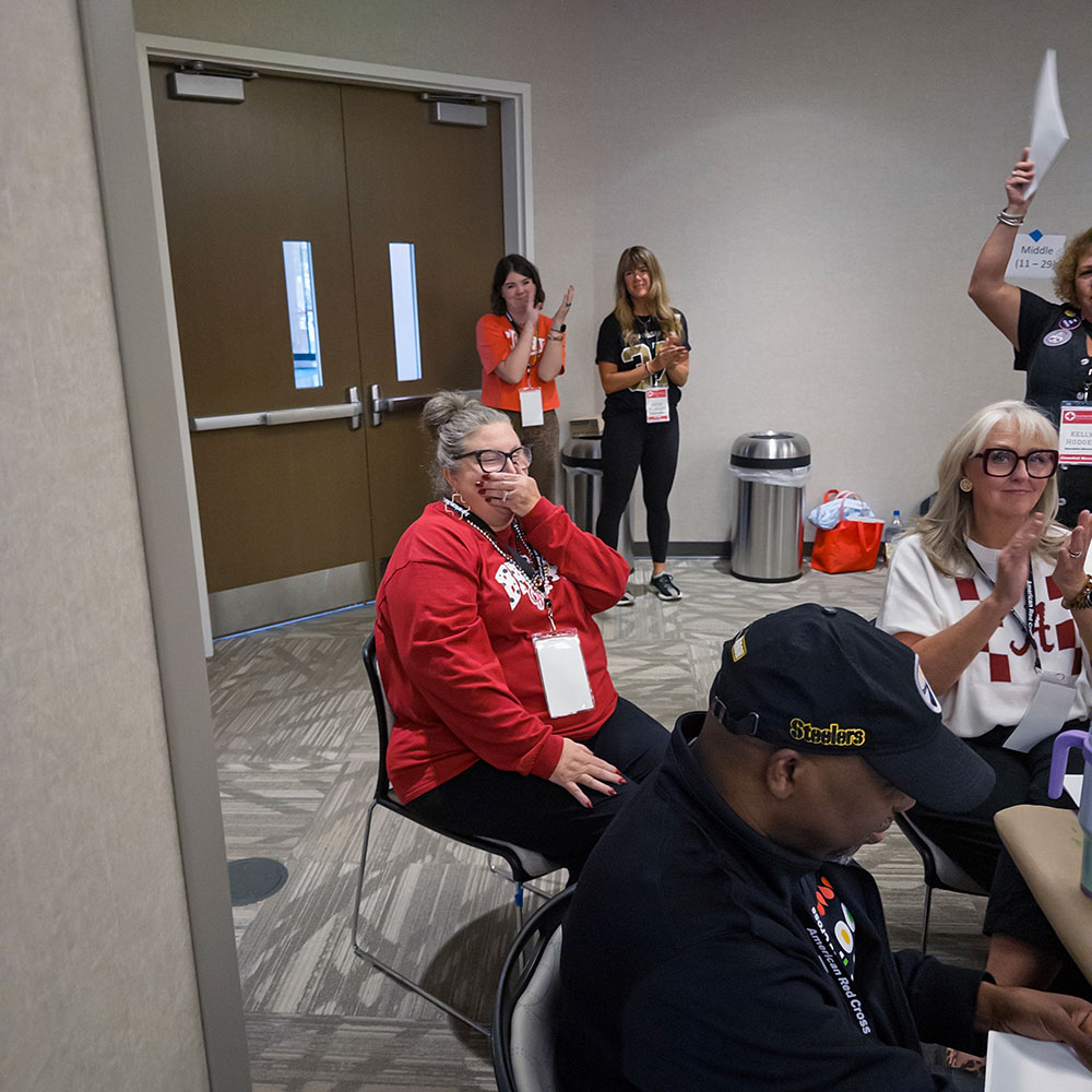 Lisa Johnson sitting in a chair with her hand covering her mouth and a room full of people clapping for her.