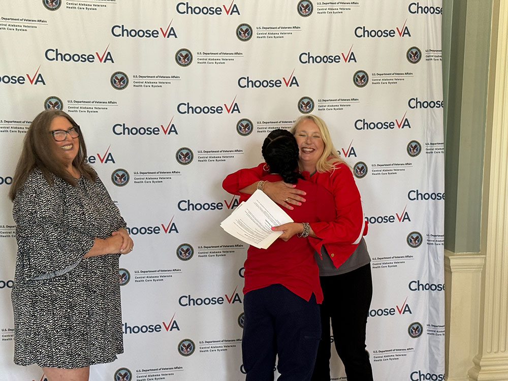 Shavonne Smith and two women stand in front of a "Choose VA" backdrop; one is hugging another while the third woman smiles and watches.