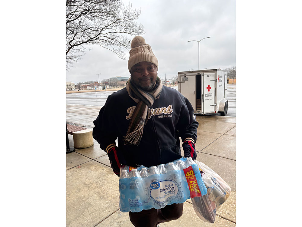 person wearing a beanie, scarf, jacket, and gloves while carrying water bottles and food from a Red Cross trailer.