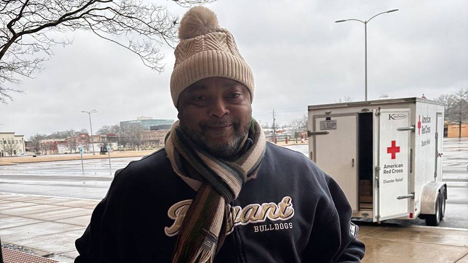 person wearing a beanie, scarf, jacket, and gloves while carrying water bottles and food from a Red Cross trailer.