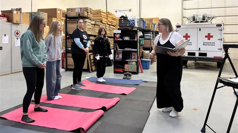 A group of four women listen attentively to an instructor in a warehouse setting with American Red Cross logos. Yoga mats are laid out on the floor.