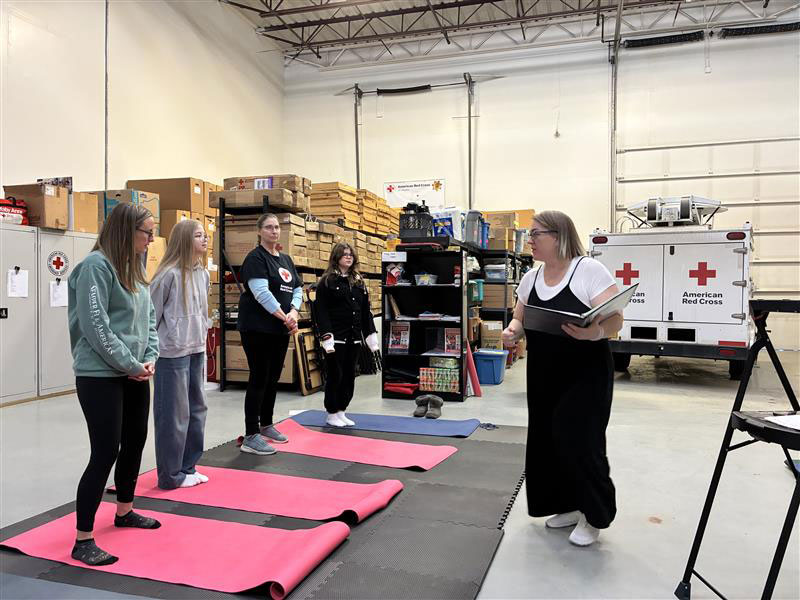A group of four women listen attentively to an instructor in a warehouse setting with American Red Cross logos. Yoga mats are laid out on the floor.